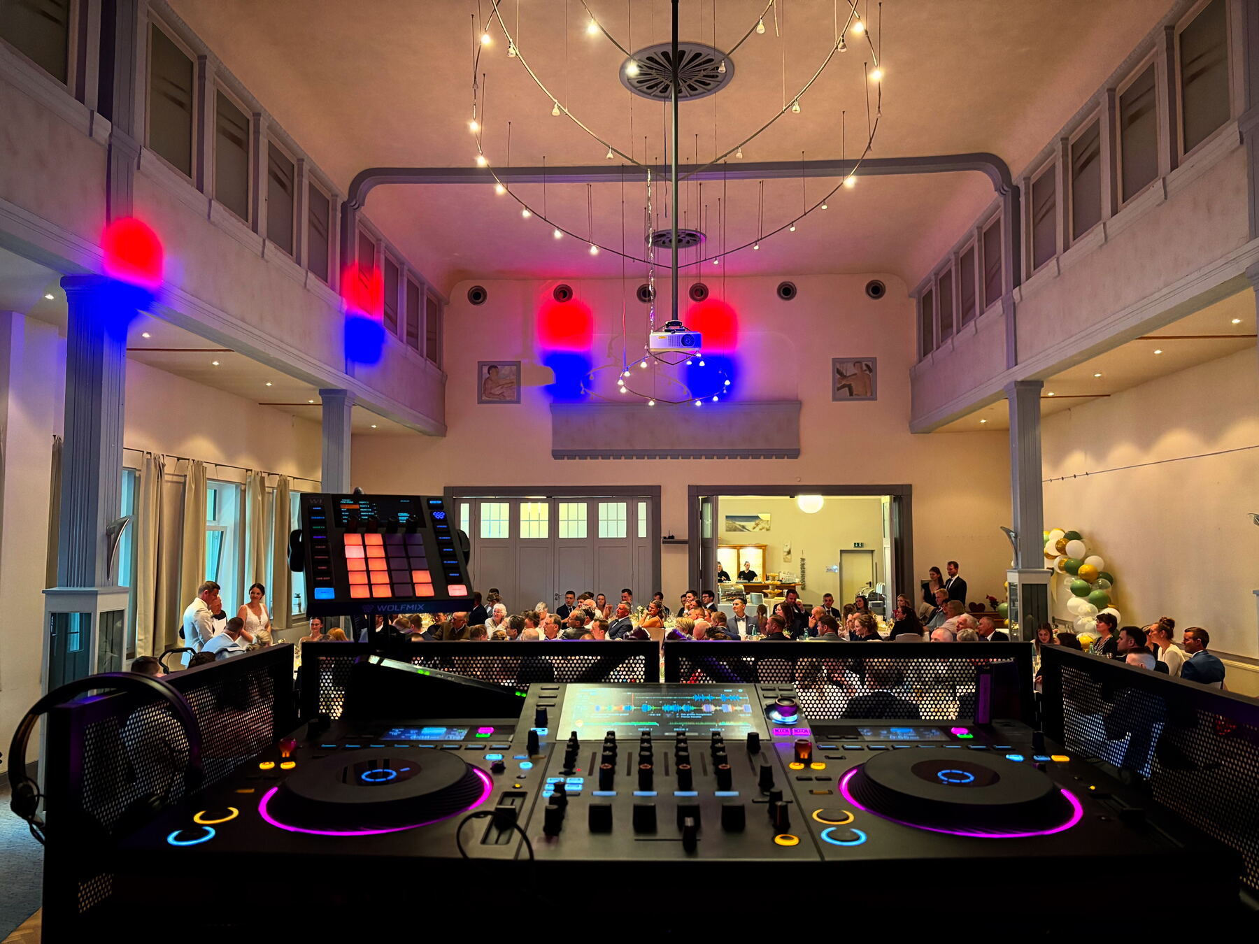 Blick hinter dem DJ-Pult auf eine volle Hochzeitsfeier mit Lichterketten
